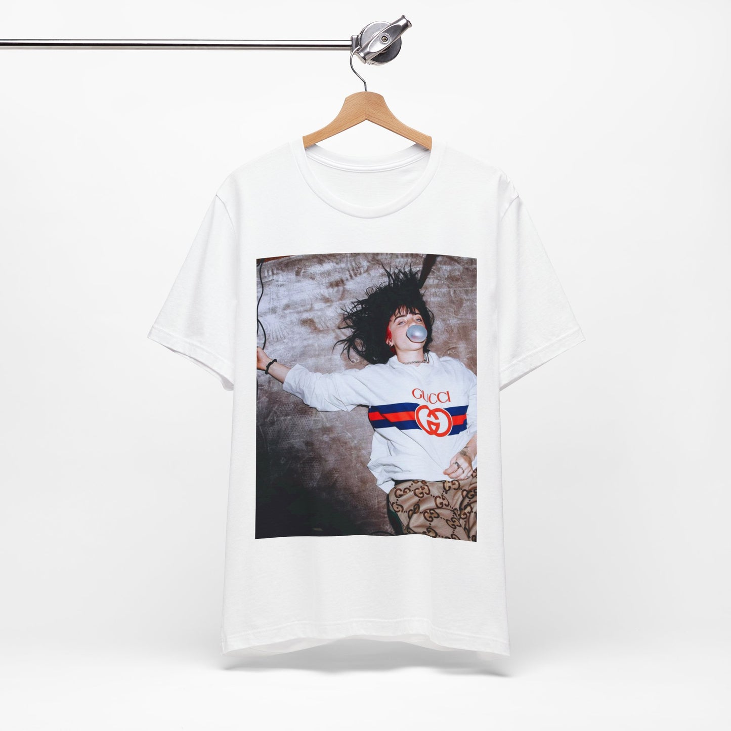 An overhead, pop-art style photo of Billie Eilish lying on the floor wearing a Gucci sweatshirt and blowing a bubblegum bubble, printed on a graphic t-shirt.