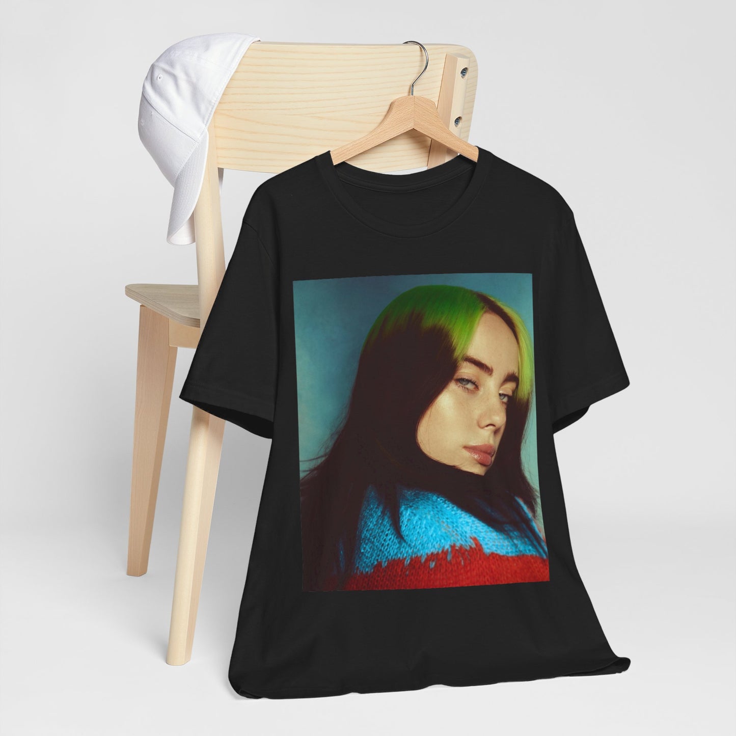 A close-up portrait of Billie Eilish with her signature green-root and black hair, wearing a blue and red sweater, printed on a graphic t-shirt.