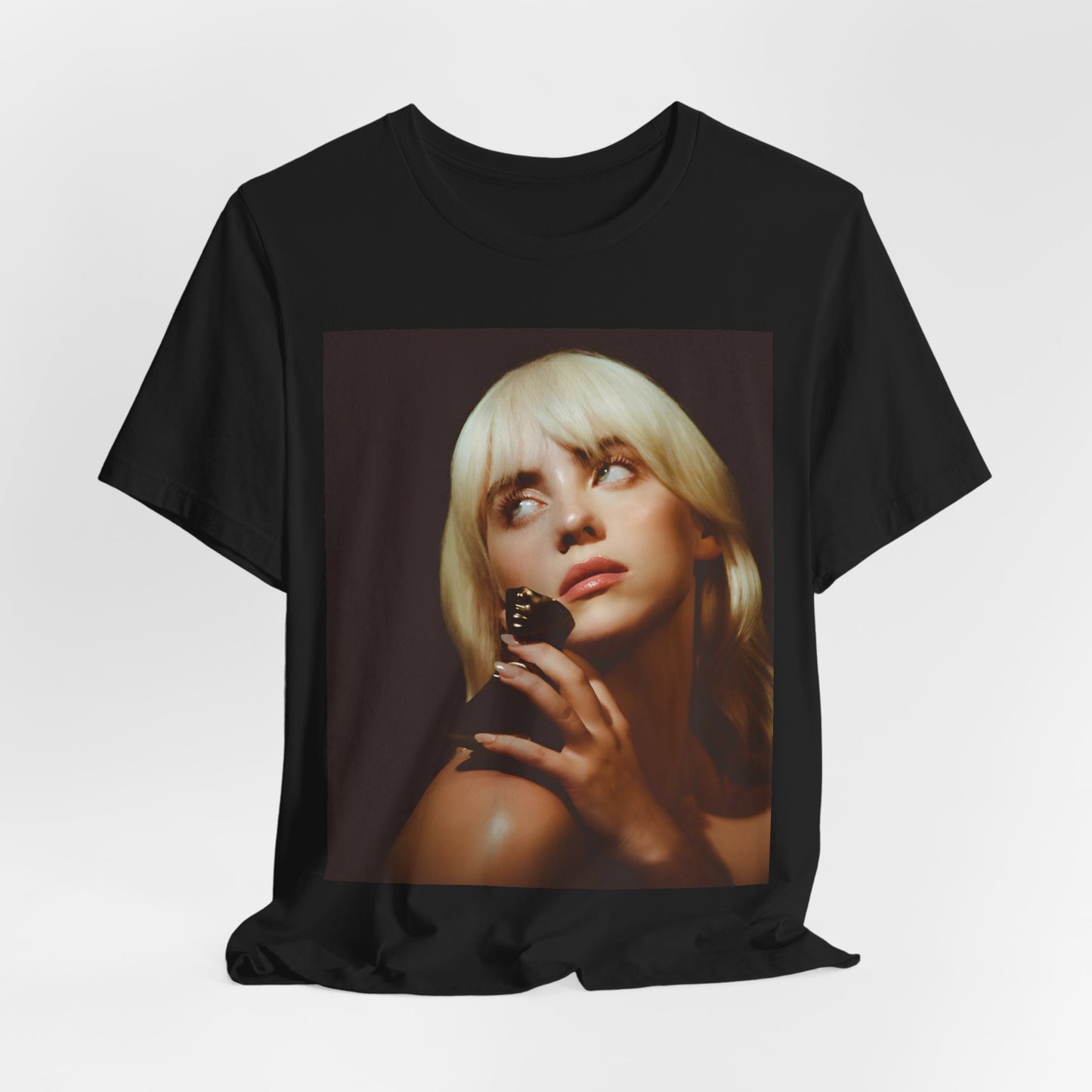 A soft, close-up portrait of Billie Eilish with dark hair looking down thoughtfully, printed on a graphic t-shirt.
