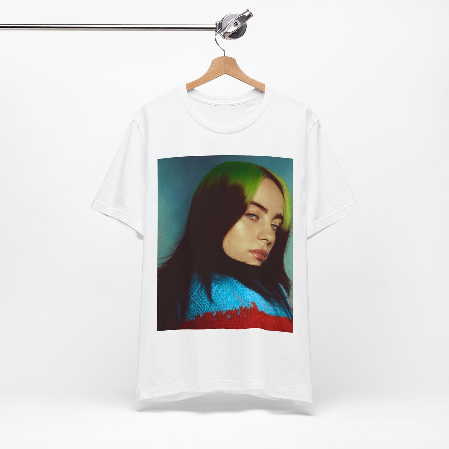 A close-up portrait of Billie Eilish with her signature green-root and black hair, wearing a blue and red sweater, printed on a graphic t-shirt.