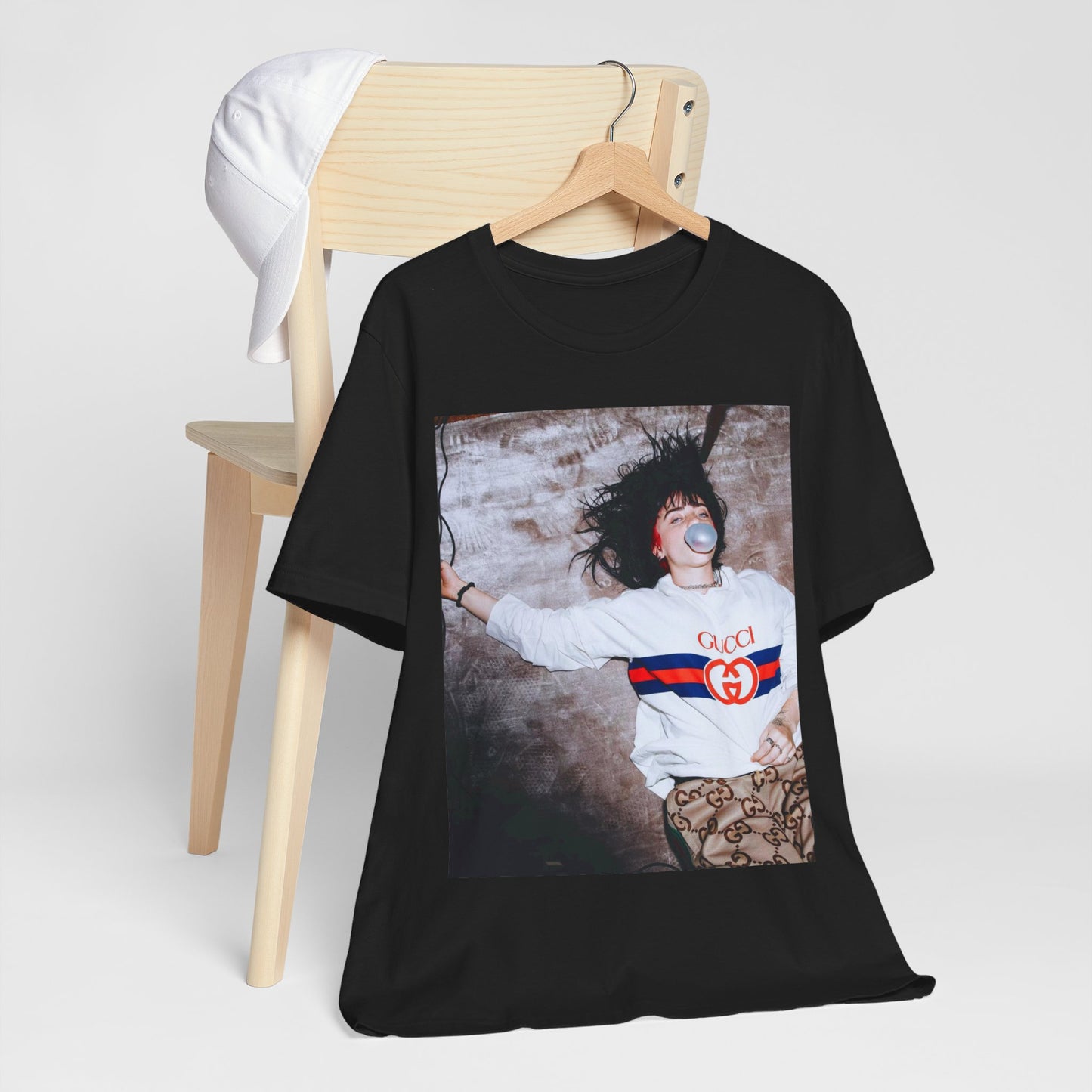 An overhead, pop-art style photo of Billie Eilish lying on the floor wearing a Gucci sweatshirt and blowing a bubblegum bubble, printed on a graphic t-shirt.