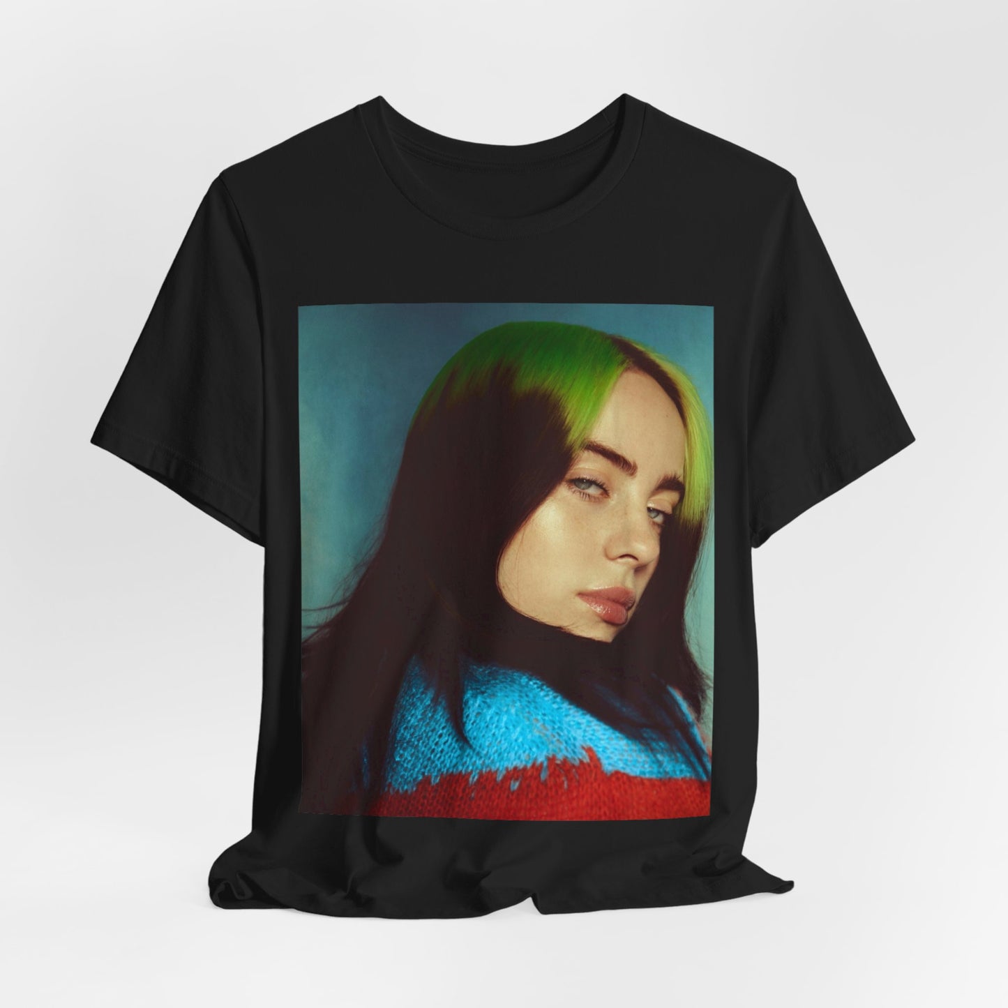 A close-up portrait of Billie Eilish with her signature green-root and black hair, wearing a blue and red sweater, printed on a graphic t-shirt.