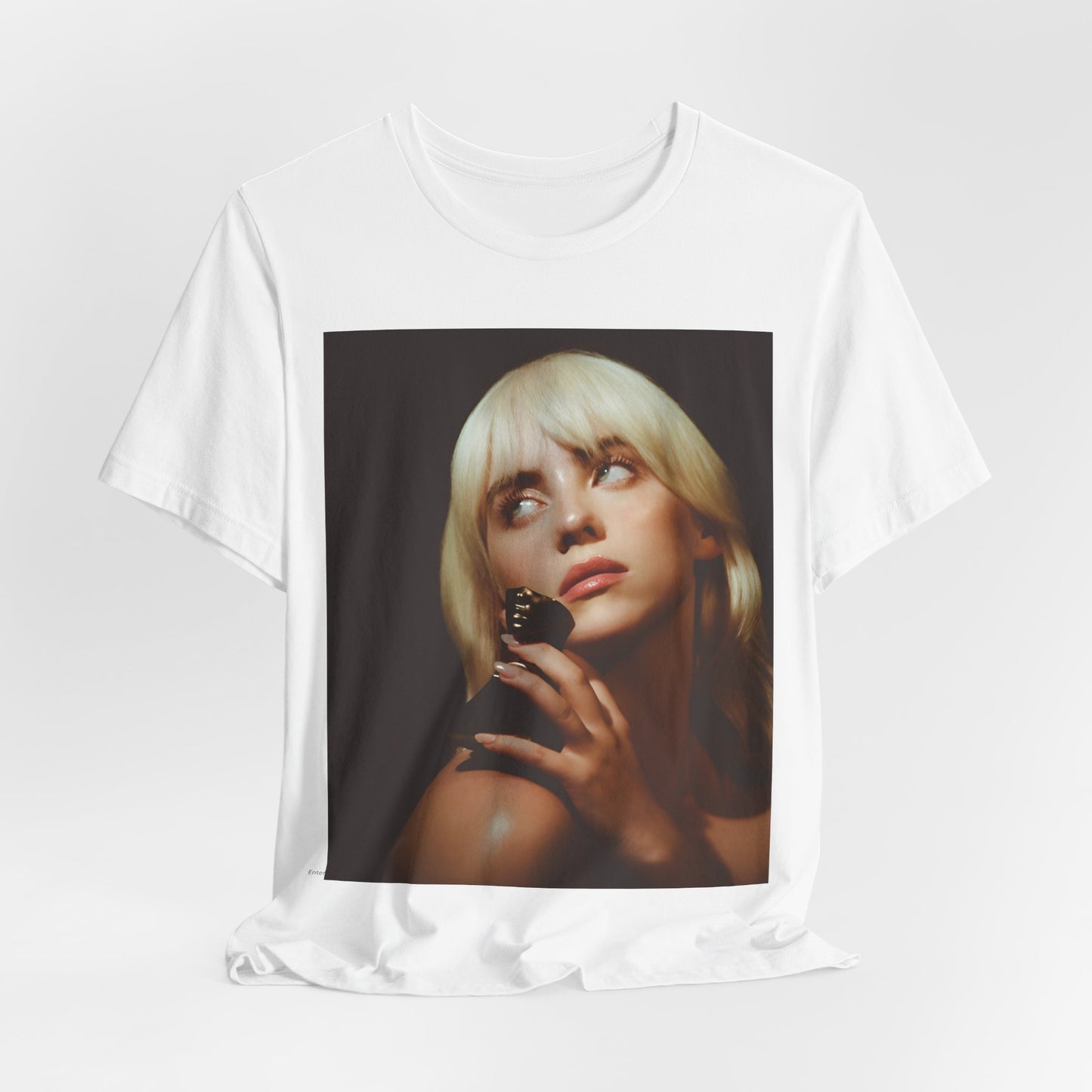 A soft, close-up portrait of Billie Eilish with dark hair looking down thoughtfully, printed on a graphic t-shirt.