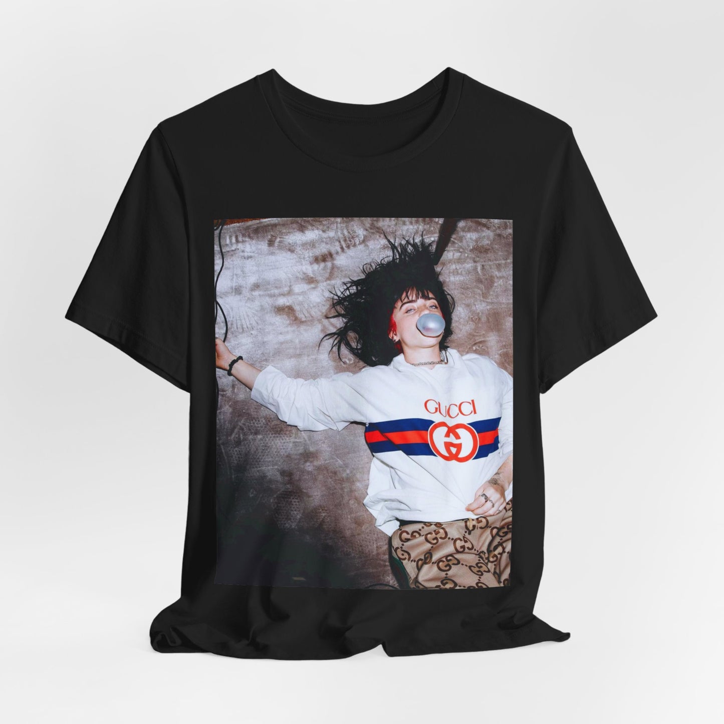 An overhead, pop-art style photo of Billie Eilish lying on the floor wearing a Gucci sweatshirt and blowing a bubblegum bubble, printed on a graphic t-shirt.