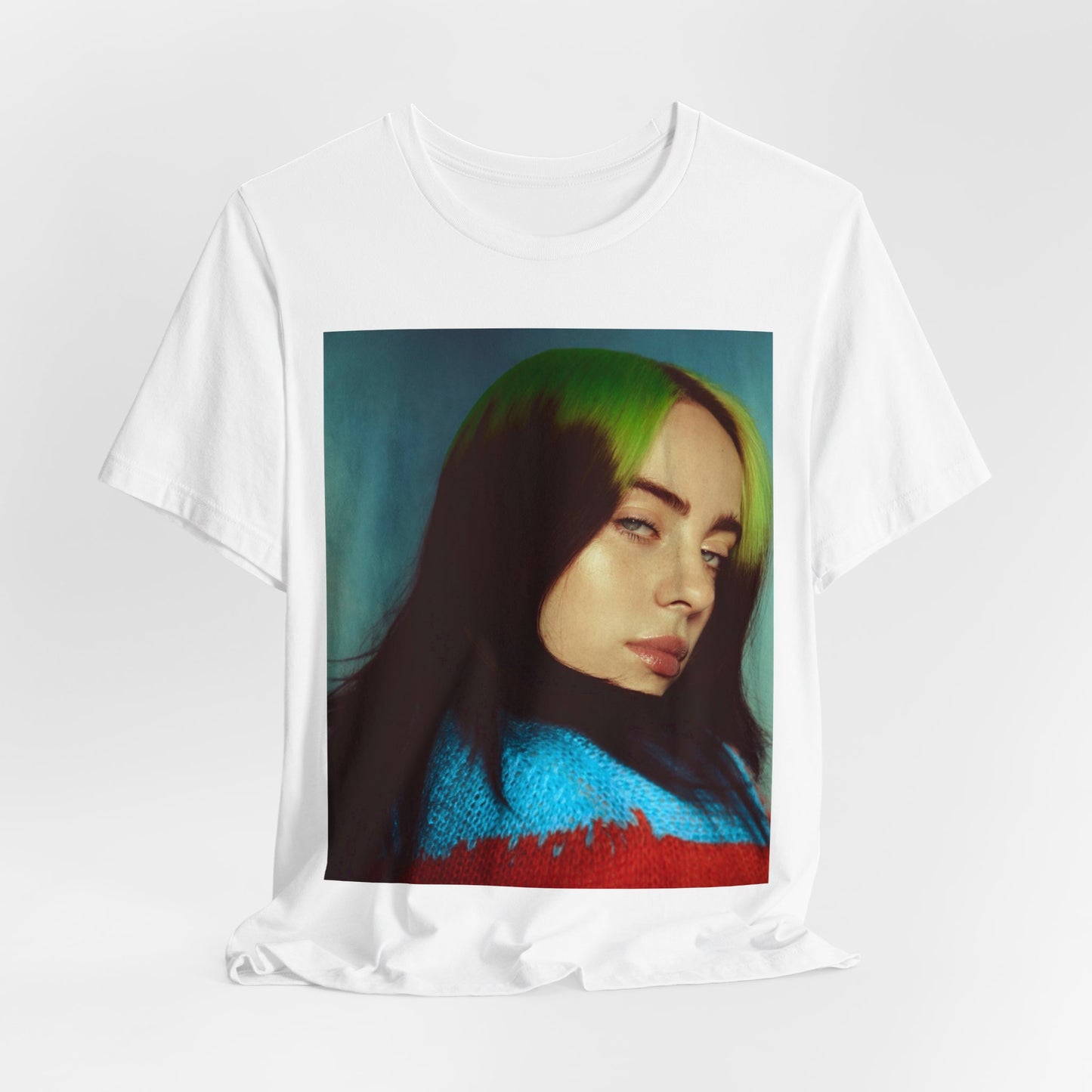 A close-up portrait of Billie Eilish with her signature green-root and black hair, wearing a blue and red sweater, printed on a graphic t-shirt.