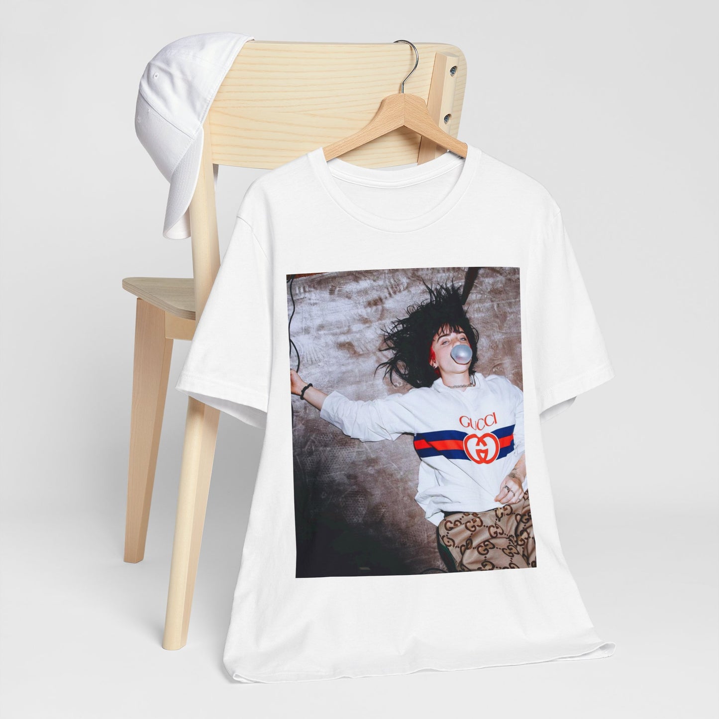 An overhead, pop-art style photo of Billie Eilish lying on the floor wearing a Gucci sweatshirt and blowing a bubblegum bubble, printed on a graphic t-shirt.
