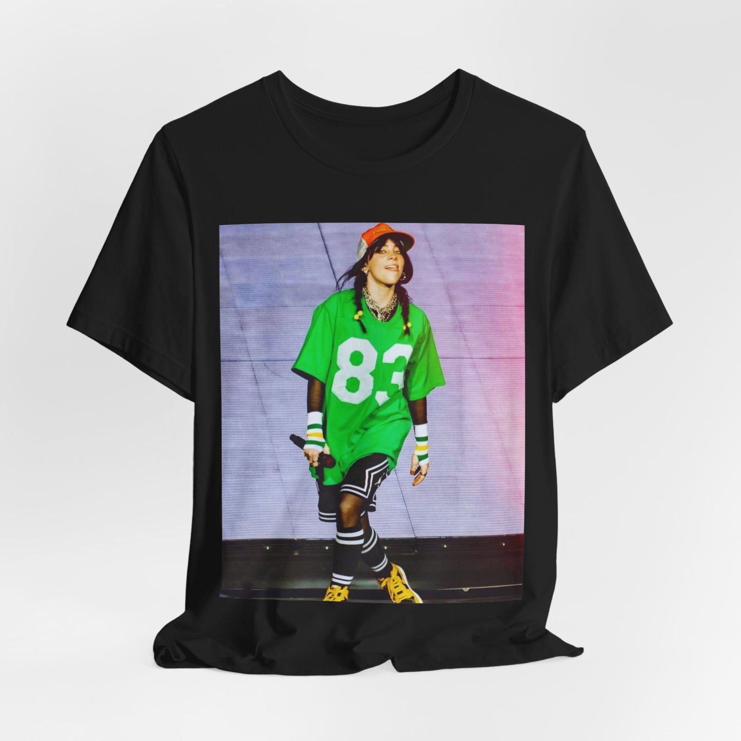 Billie Eilish performing on stage wearing a large green basketball jersey with the number 83 and an orange backwards cap.