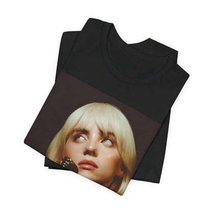 A soft, close-up portrait of Billie Eilish with dark hair looking down thoughtfully, printed on a graphic t-shirt.