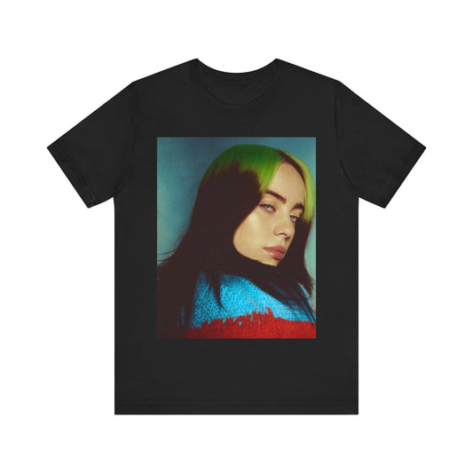 A close-up portrait of Billie Eilish with her signature green-root and black hair, wearing a blue and red sweater, printed on a graphic t-shirt.