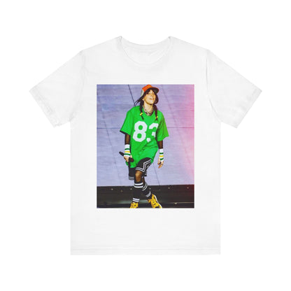 Billie Eilish performing on stage wearing a large green basketball jersey with the number 83 and an orange backwards cap.