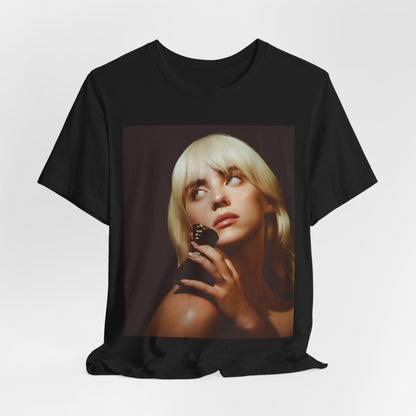 A soft, close-up portrait of Billie Eilish with dark hair looking down thoughtfully, printed on a graphic t-shirt.