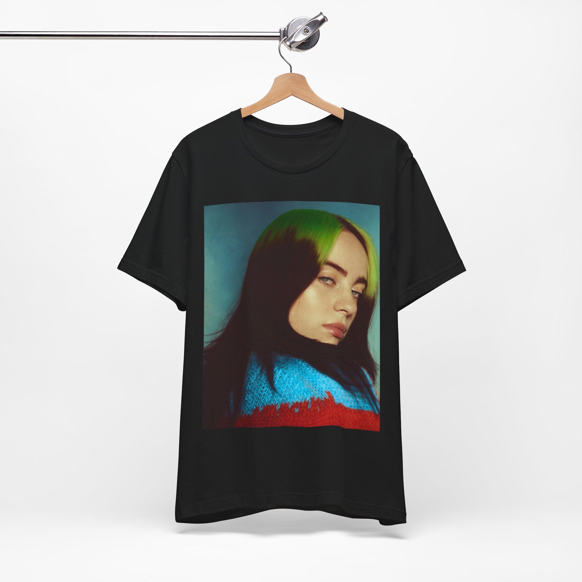 A close-up portrait of Billie Eilish with her signature green-root and black hair, wearing a blue and red sweater, printed on a graphic t-shirt.