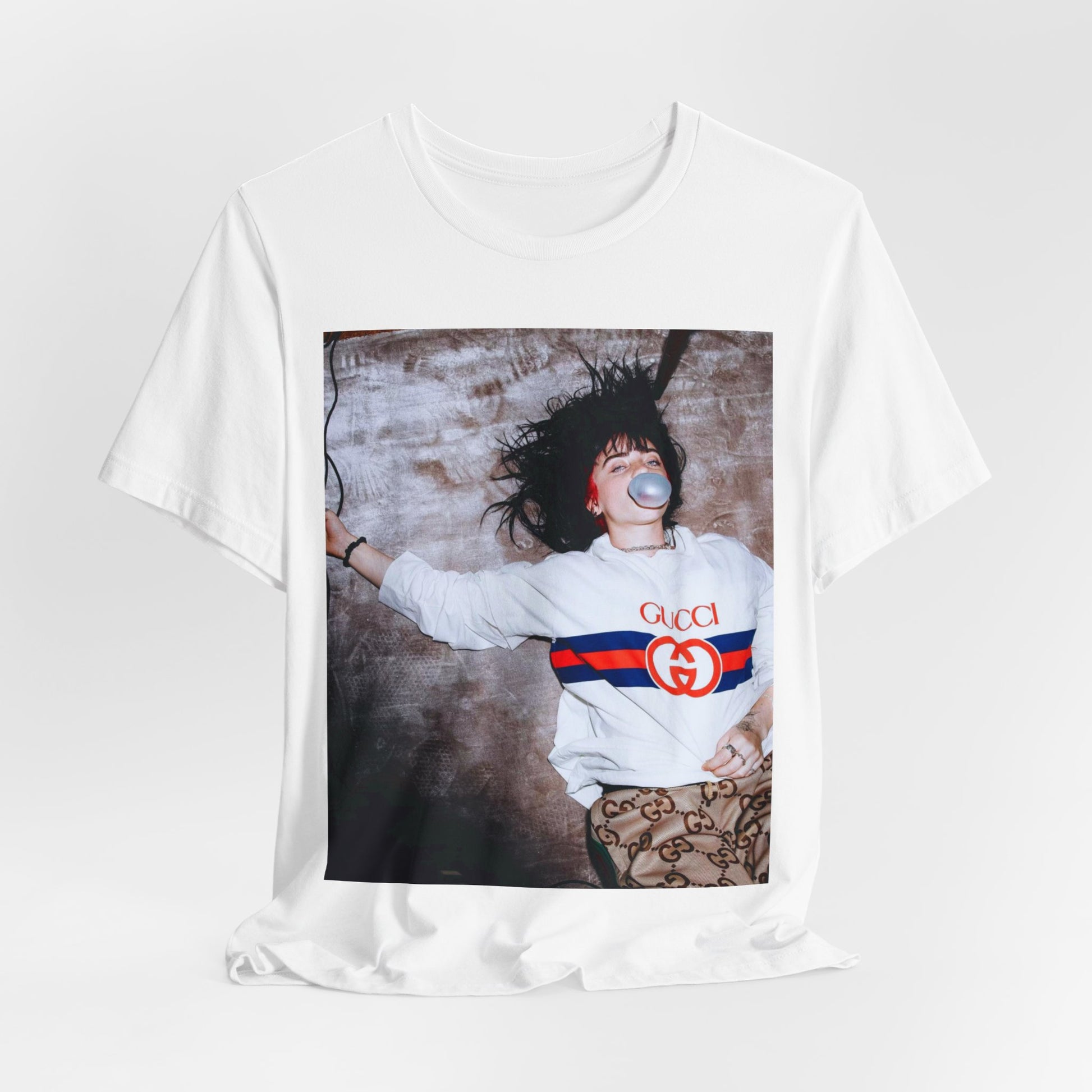 An overhead, pop-art style photo of Billie Eilish lying on the floor wearing a Gucci sweatshirt and blowing a bubblegum bubble, printed on a graphic t-shirt.