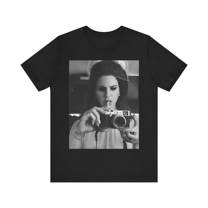 Black and white photo of Lana Del Rey holding a vintage camera and a cigarette to her lips, printed on a graphic tee.