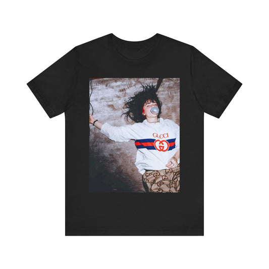 An overhead, pop-art style photo of Billie Eilish lying on the floor wearing a Gucci sweatshirt and blowing a bubblegum bubble, printed on a graphic t-shirt.
